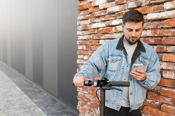 Man wearing a denim jacket on electric scooter uses smartphone against the background of brick wall. Person uses smartphone to search for geolocation. 