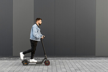 Attractive man in a denim jacket riding electric kick scooter on a grey background of a street building. Driving an ecological mode of transport.