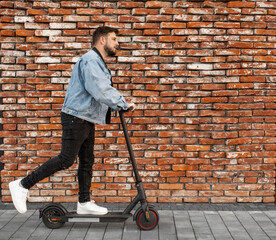 Attractive man in a denim jacket riding electric kick scooter at  brick wall background. Driving an ecological mode of transport.