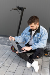 Young and handsome man with a beard sits on an electric scooter and works at his laptop against the background of a gray wall on a city street. Freelance work outside the home. 