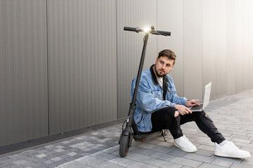 Young and handsome man with a beard sits on an electric scooter and works at his laptop against the background of a gray wall on a city street. Freelance work outside the home. 