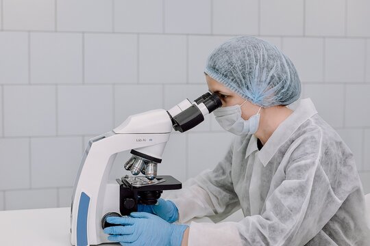 Female scientist in protective clothing and medical mask using a laboratory microscope for research in a modern clinical setting. Horizontal image with copy space.