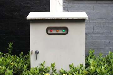 Outdoor electrical control box with indicator lights (red, orange, green) surrounded by lush green...