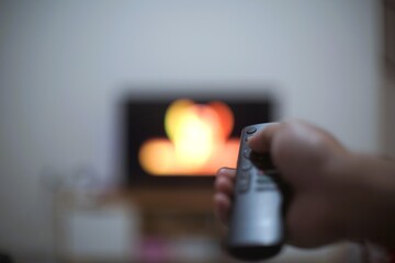 Close-up of a hand holding a remote control pointing towards a blurred TV screen with colorful bokeh lights in a dim room.