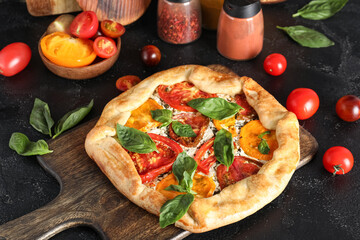 Wooden board of tasty tomato galette with basil on black background