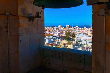 Scenic view of Cadiz city framed by church tower opening, with vibrant rooftops stretching to the sea under a clear blue sky. Historic bell structure adds depth to this elevated urban panorama