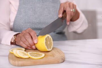 Senior woman cutting lemon at white marble table indoors, closeup