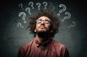 Man with curly hair and glasses looks up with thoughtful expression, surrounded by chalk question marks on dark background