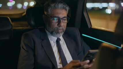Senior Indian businessman in suit and glasses scrolling, typing and reading message on smartphone while riding in backseat of car at night - Powered by Adobe
