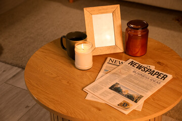 Newspapers with burning candle, cup and blank frame on table in dark room