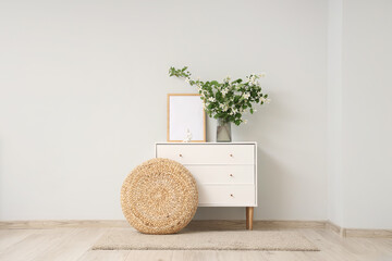 Vase of blooming jasmine flowers with frame on commode and wicker ottoman near grey wall in living...