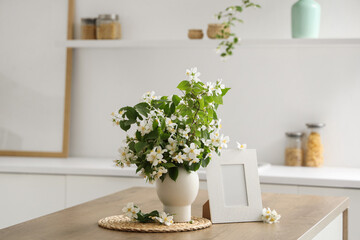 Vase of blooming jasmine flowers with blank frame on table in kitchen