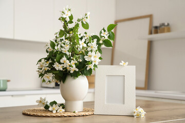 Vase of blooming jasmine flowers with blank frame on table in kitchen