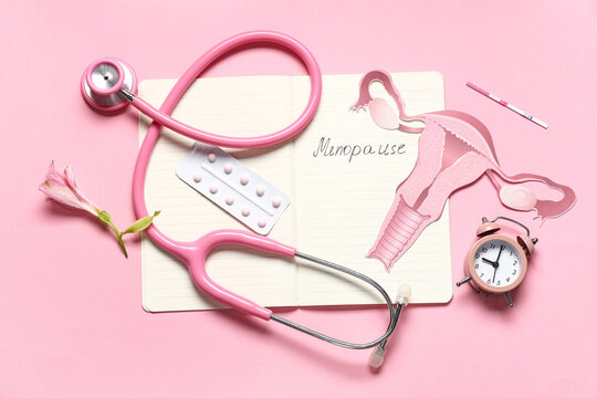 Notebook with word MENOPAUSE, pills, stethoscope and paper uterus on pink background