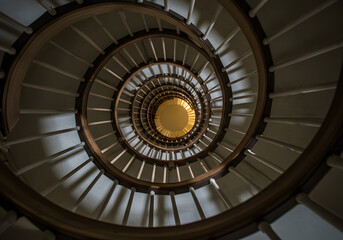 Obraz premium A low-angle view of a spiral staircase with white steps and a wooden railing, leading upwards to a bright light.