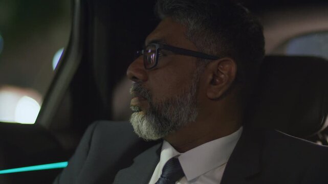 Senior grey-haired businessman in formal suit and glasses looking through car window at blurred city lights during nighttime ride - Powered by Adobe