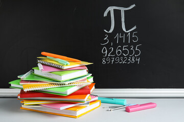 Number Pi written on black chalkboard with notebooks and stationery on table in classroom
