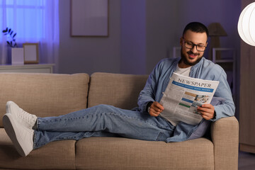 Handsome man with eyeglasses reading newspaper on sofa at home in evening