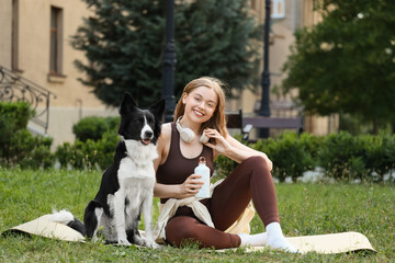 Sporty young woman with bottle of water and cute Border Collie dog sitting on yoga mat in park