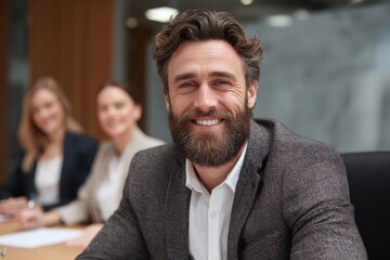 A smiling young Caucasian businessman in formal attire is seated at a table with female colleagues chatting behind him