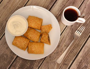 A plate with several pancakes stuffed with minced meat, a cup of sour cream, a fork and hot black tea on a wooden table.