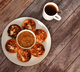 Fresh cheesecakes with raisins in a round white plate, a cup of apple jam in the center and hot tea on a wooden table.