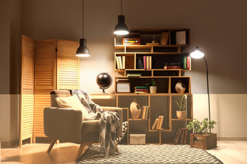 Interior of stylish living room with grey armchair, glowing lamps and bookshelves at evening