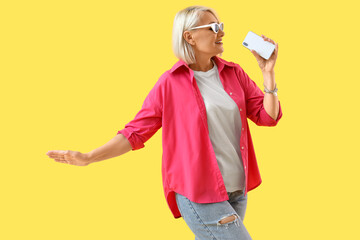 Mature woman in sunglasses with mobile phone singing on yellow background