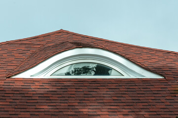 The exterior of a red asphalt peaked roof with an eyebrow window. The curved window has a vinyl frame with a clear fixed glass pane. The unique shape of the window resembles an eyebrow with its curve. © Dolores  Harvey
