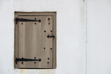 The exterior white cement wall of a building with a small, closed wooden door. The hinges on the door are rusty strap hinges. The latch is curved in shape with a hook and peg form. The door is wooden.