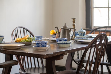 The dining room of a vintage building with white walls, single hung window, long wooden table, bow back chairs, metal kettle, candlestick holder, blue dishes, and fruit. The tableware is vintage.