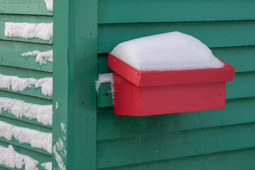 The exterior corner of two wooden walls of a cottage with a small red wooden mailbox on the right side. The left wall has fresh white snow stuck to the rough horizontal boards. Snow is on the lid.