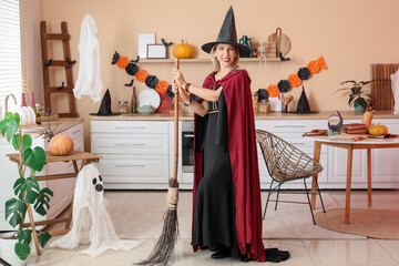 Happy mature woman dressed as witch with broom in decorated kitchen. Halloween celebration