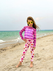 Stylish young girl posing confidently on the beach, wearing colorful leggings with star patterns and a pink tie-dye top. She sports yellow flower-shaped sunglasses. Wind in her hair