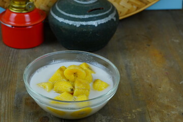 A glass bowl of kolak pisang or Indonesian banana compote in coconut milk, with a red oil lamp and a traditional clay pot in the background.