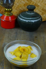 A glass bowl of kolak pisang or Indonesian banana compote in coconut milk, with a red oil lamp and a traditional clay pot in the background.
