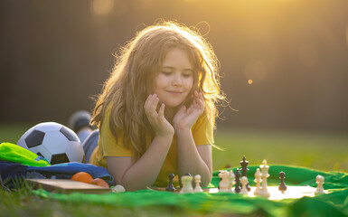 Blonde cute kid play chess outdoors. Kid thinking during a chess game. Child playing chess on the...