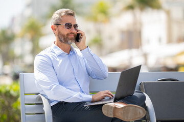 Confident businessman sitting on a bench with a laptop and phone in park. Successful businessman....