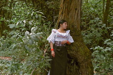 Indigenous woman wearing traditional clothes leaning against a tree in the forest