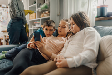 Children watching smartphone while mother cleaning living room