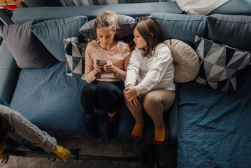 Mother vacuuming floor while daughters relaxing on sofa using smartphone