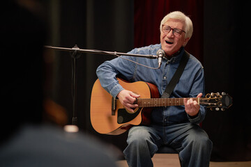 Senior man singing and playing acoustic guitar on stage during talent competition, sitting in front of microphone, performing passionately for audience