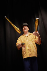 Senior Caucasian man juggling golden clubs on stage during talent competition, wearing hat and...