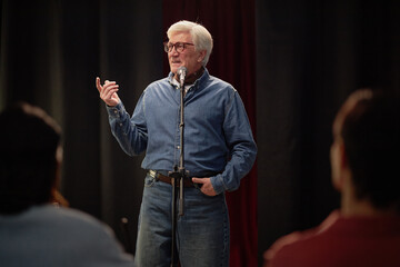 Senior Caucasian man standing on stage speaking into microphone during talent competition,...
