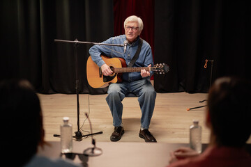 Senior Caucasian man sitting on stage playing acoustic guitar and singing into microphone during talent competition performance with two judges watching in foreground