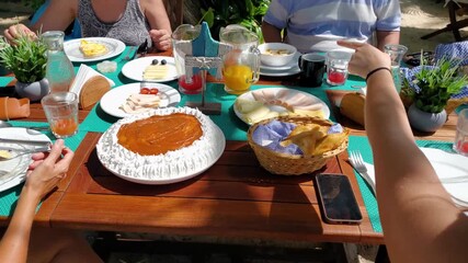 Eevated view Family around a wooden table laden with various dishes, including a large cake-like dessert, pastries, scrambled eggs, and fruit and empanadas - Powered by Adobe