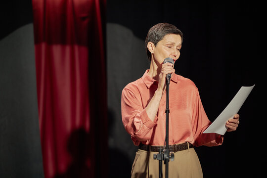 Middle aged Caucasian woman standing on stage holding microphone and reading from paper during talent competition, focused expression, short hair, stage curtain in background