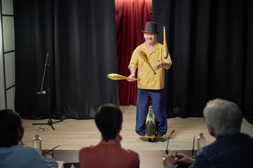 Caucasian man juggling clubs while balancing on unicycle on stage during talent competition, smiling and performing in front of three seated judges holding pens © Seventyfour