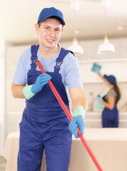 Portrait of smiling young professional worker of cleaning service in blue uniform and rubber gloves...