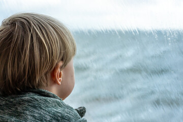 Ein kleiner Junge guckt sehnsüchtig durch eine nasse Fensterscheibe bei Regenwetter